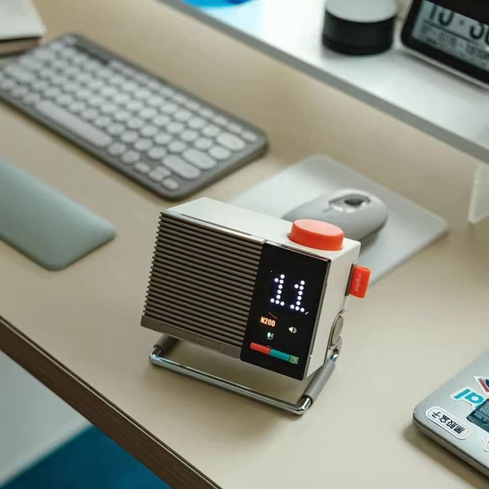 Boxy retro-futuristic desk speaker with red push-button and dot-matrix display, on a workspace