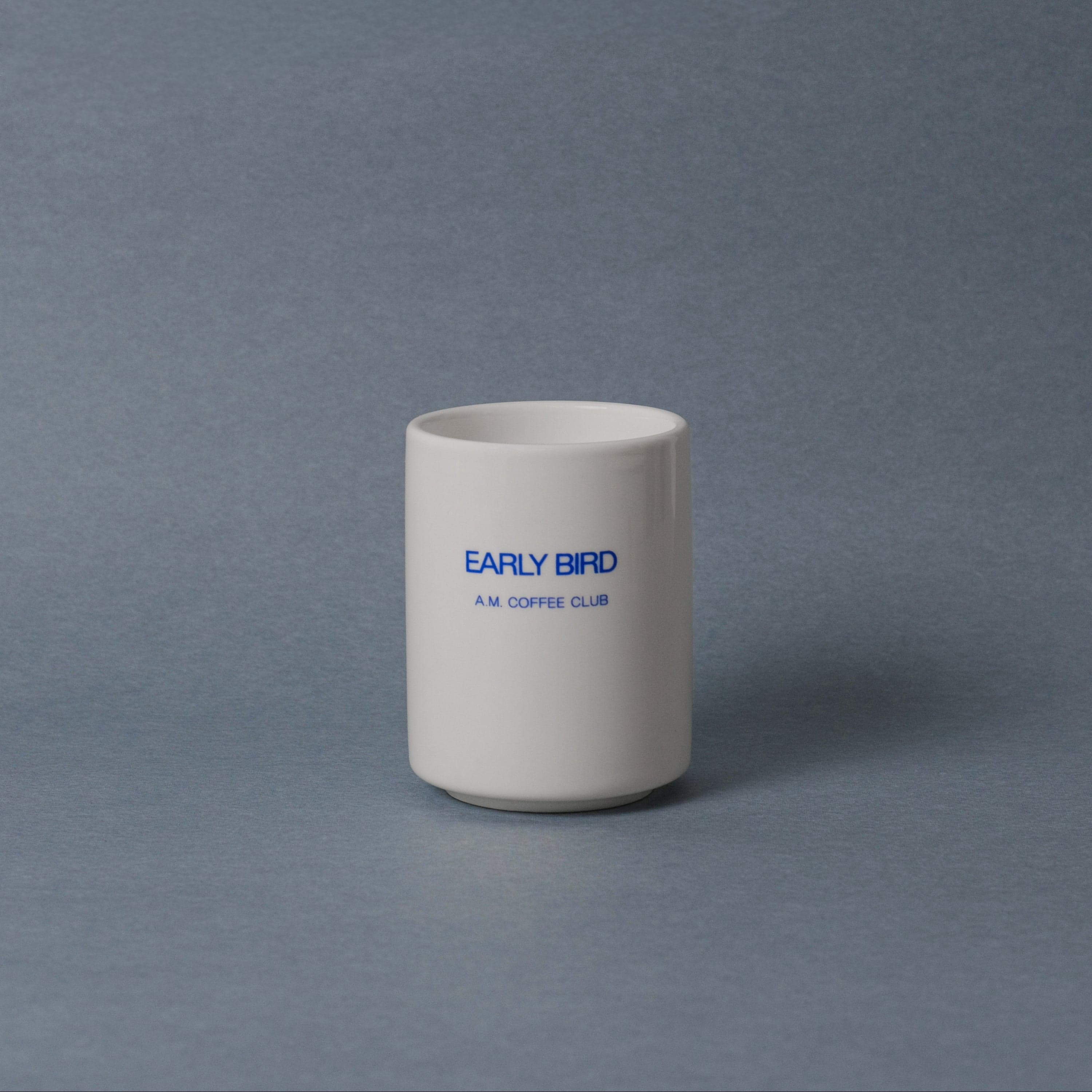 Small white ceramic cup printed 'EARLY BIRD A.M. COFFEE CLUB' in cobalt blue on a slate-grey seamless