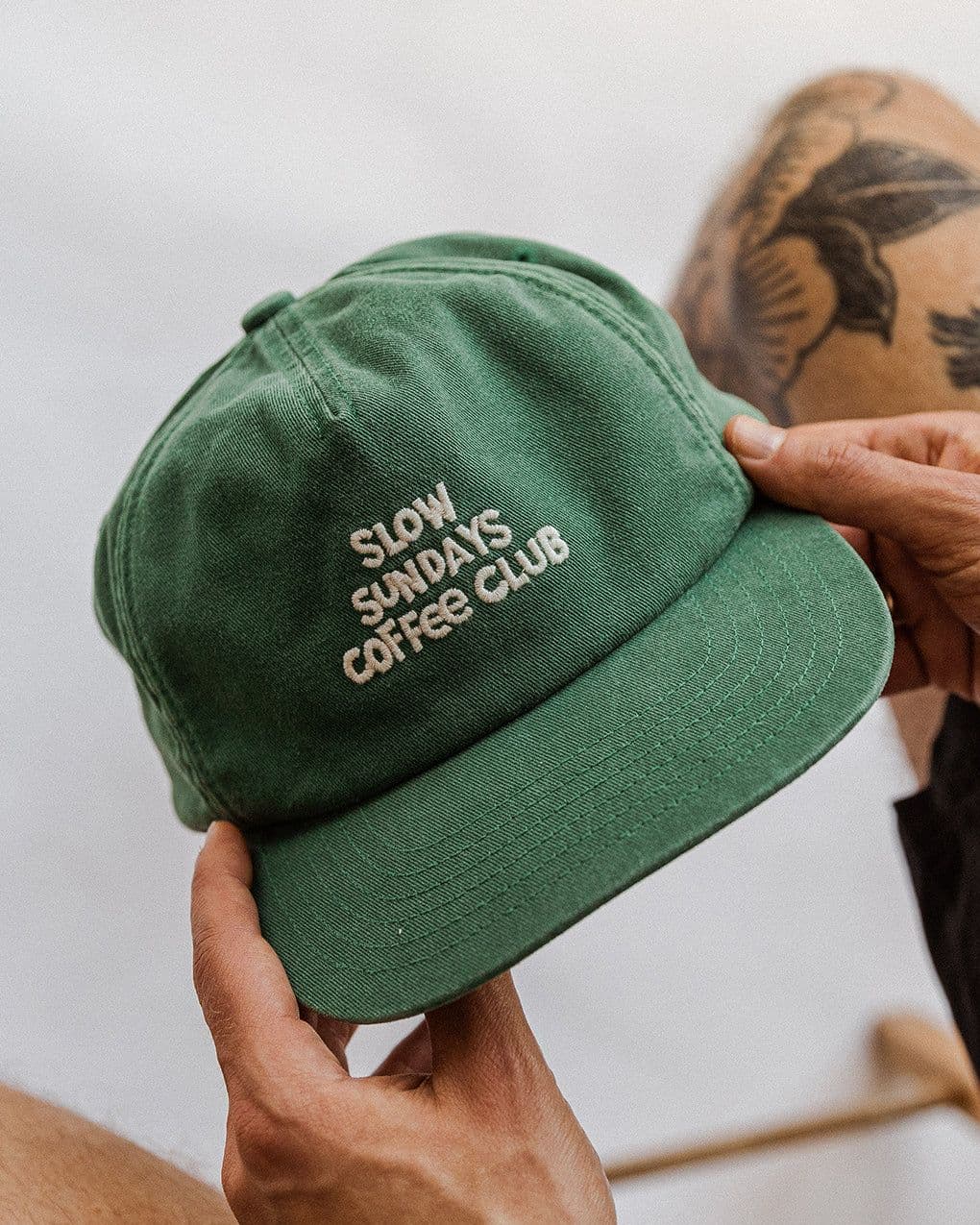 Hands holding a forest-green five-panel cap embroidered "Slow Sundays Coffee Club" in cream, against a soft out-of-focus backdrop