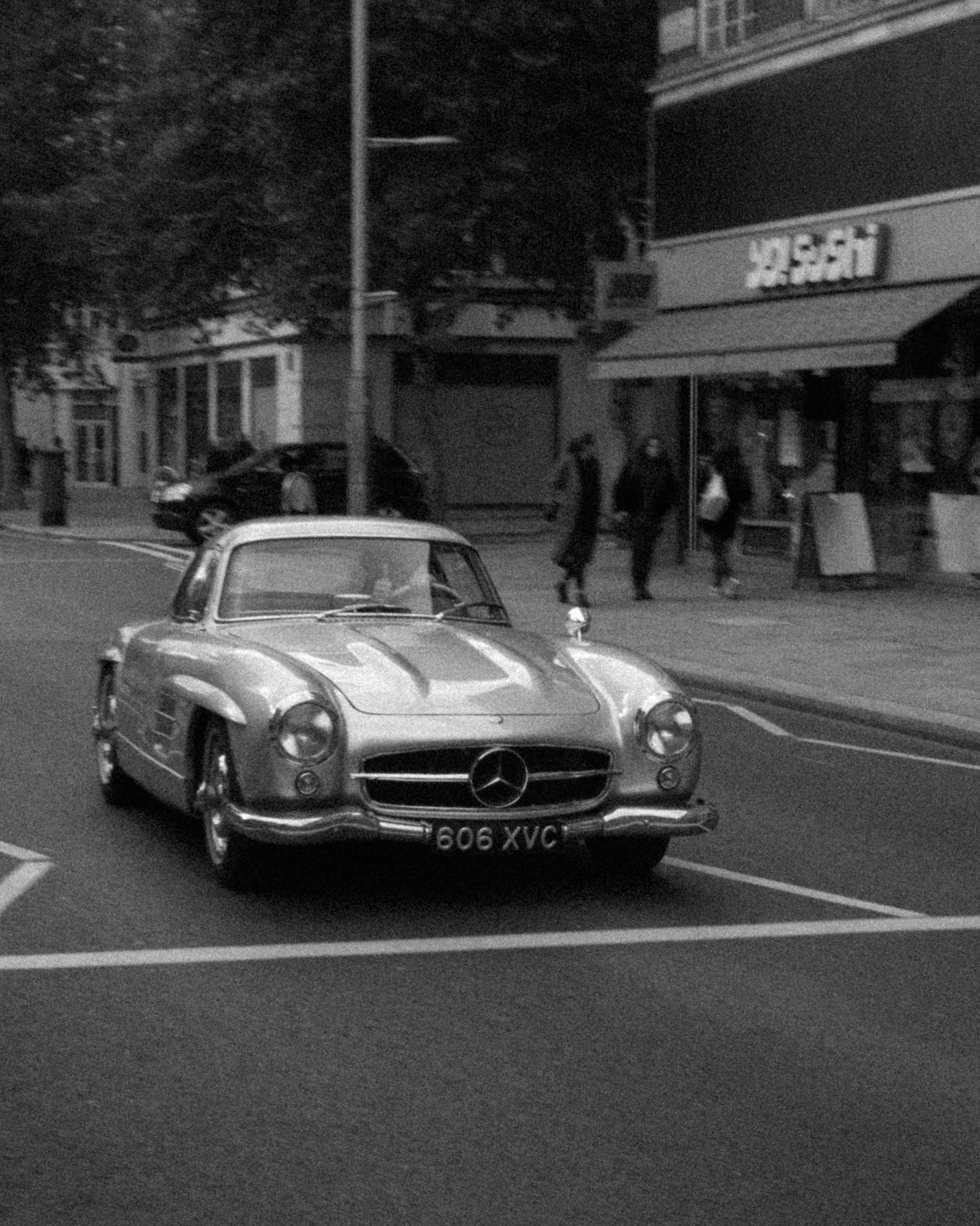 Black-and-white street photo of a silver Mercedes 300SL Gullwing driving past shopfronts and pedestrians on a city road